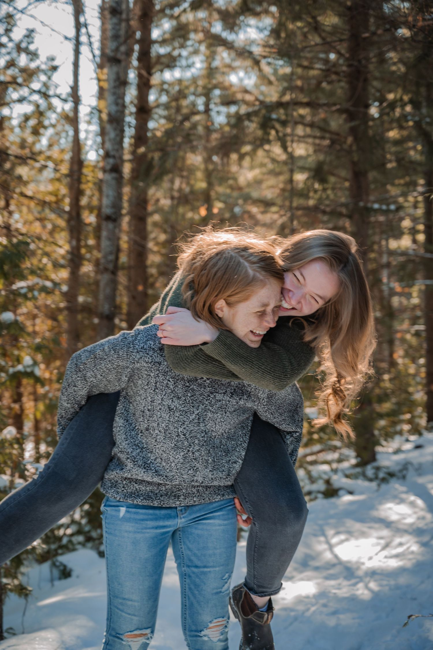 A playful couple in grey sweaters and jeans embrace while enjoying a snowy winter forest scene.