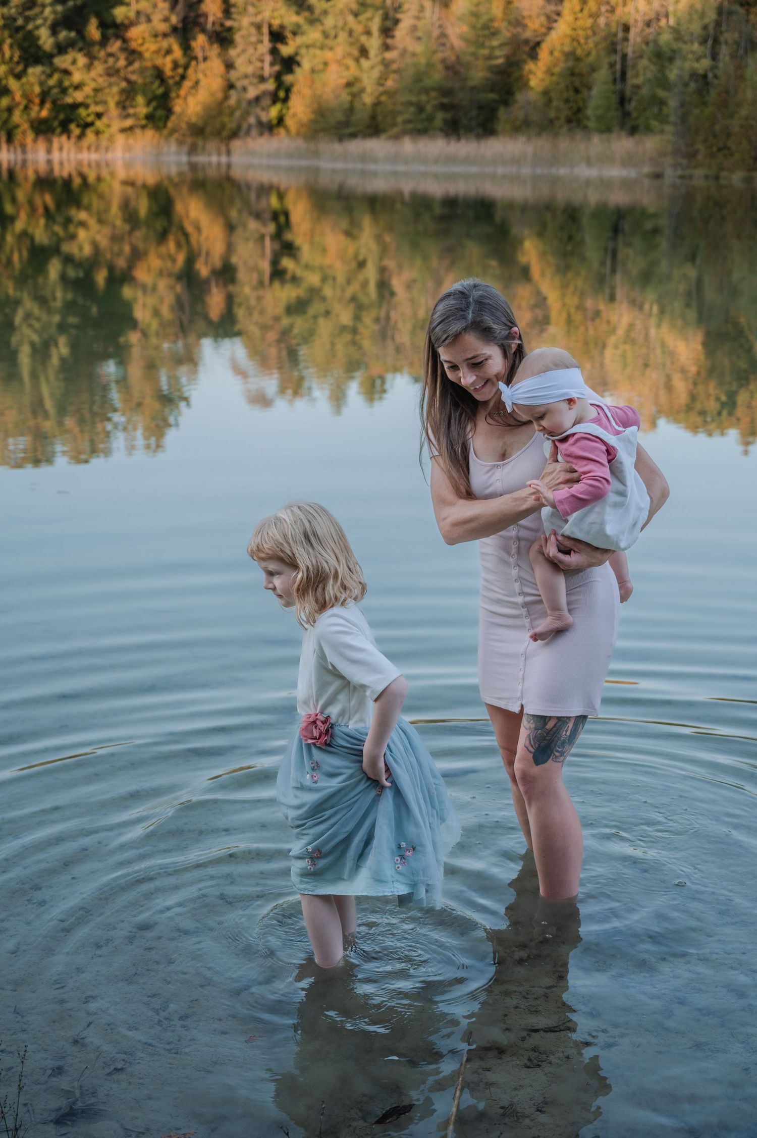 A mother holding a baby and a young blonde girl wade in a calm reflective lake surrounded by autumn trees.