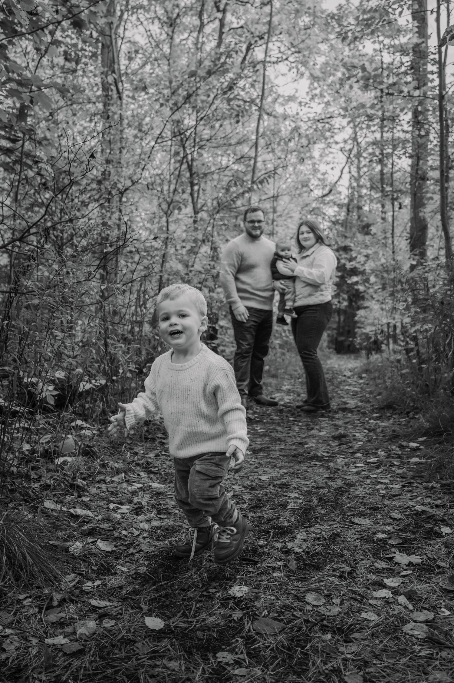 Black and white photo of a toddler running on a forest path while parents walk behind holding a baby.