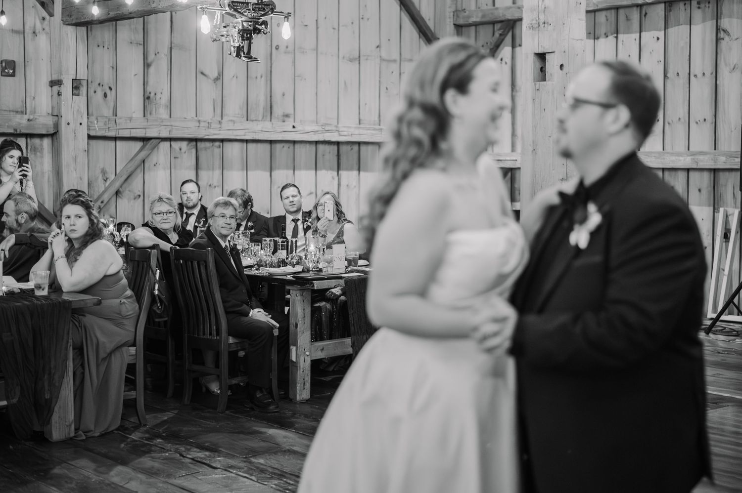 Bride and groom share first dance in rustic barn wedding venue while guests watch from decorated tables.