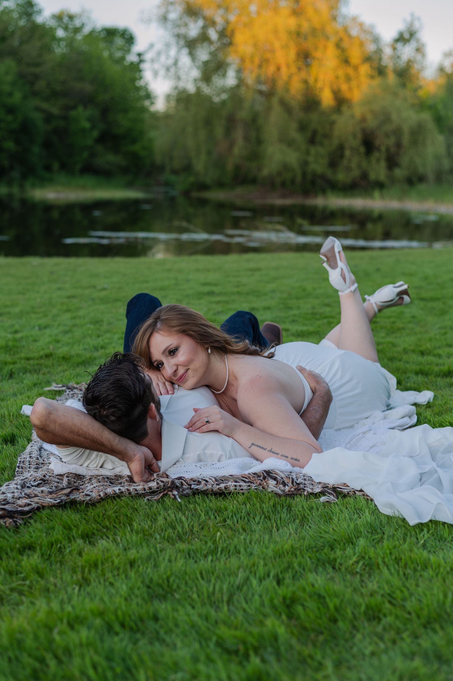 Couple in wedding attire lying on a blanket in lush green park near a pond, sharing a romantic moment.