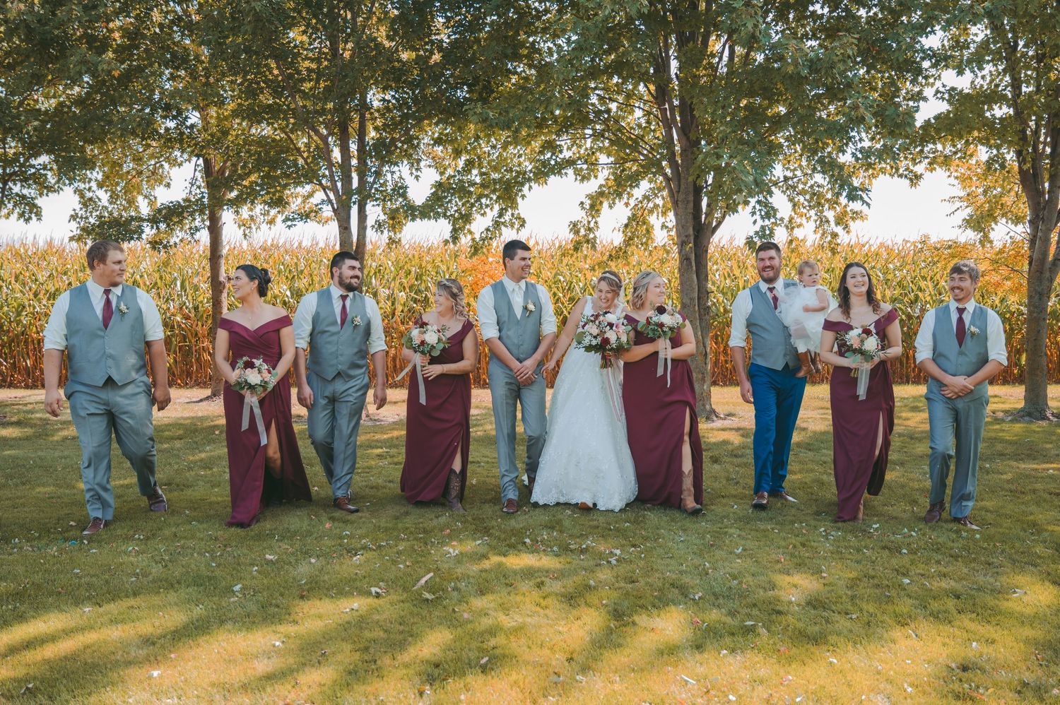 Wedding party in burgundy attire poses together on a sunny autumn day with trees and golden light in background.