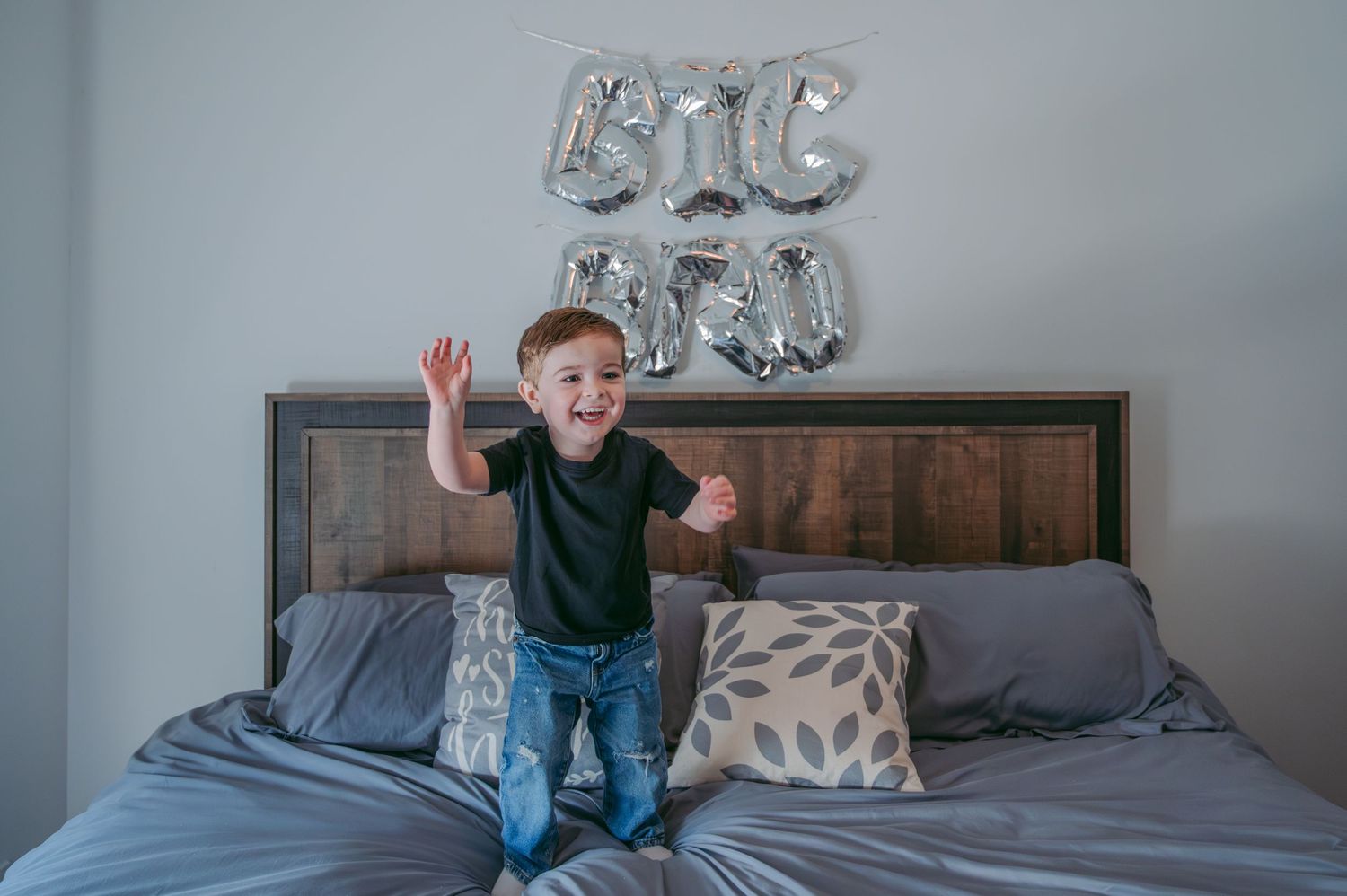 A child jumps playfully on a bed with silver balloon decorations in the background.
