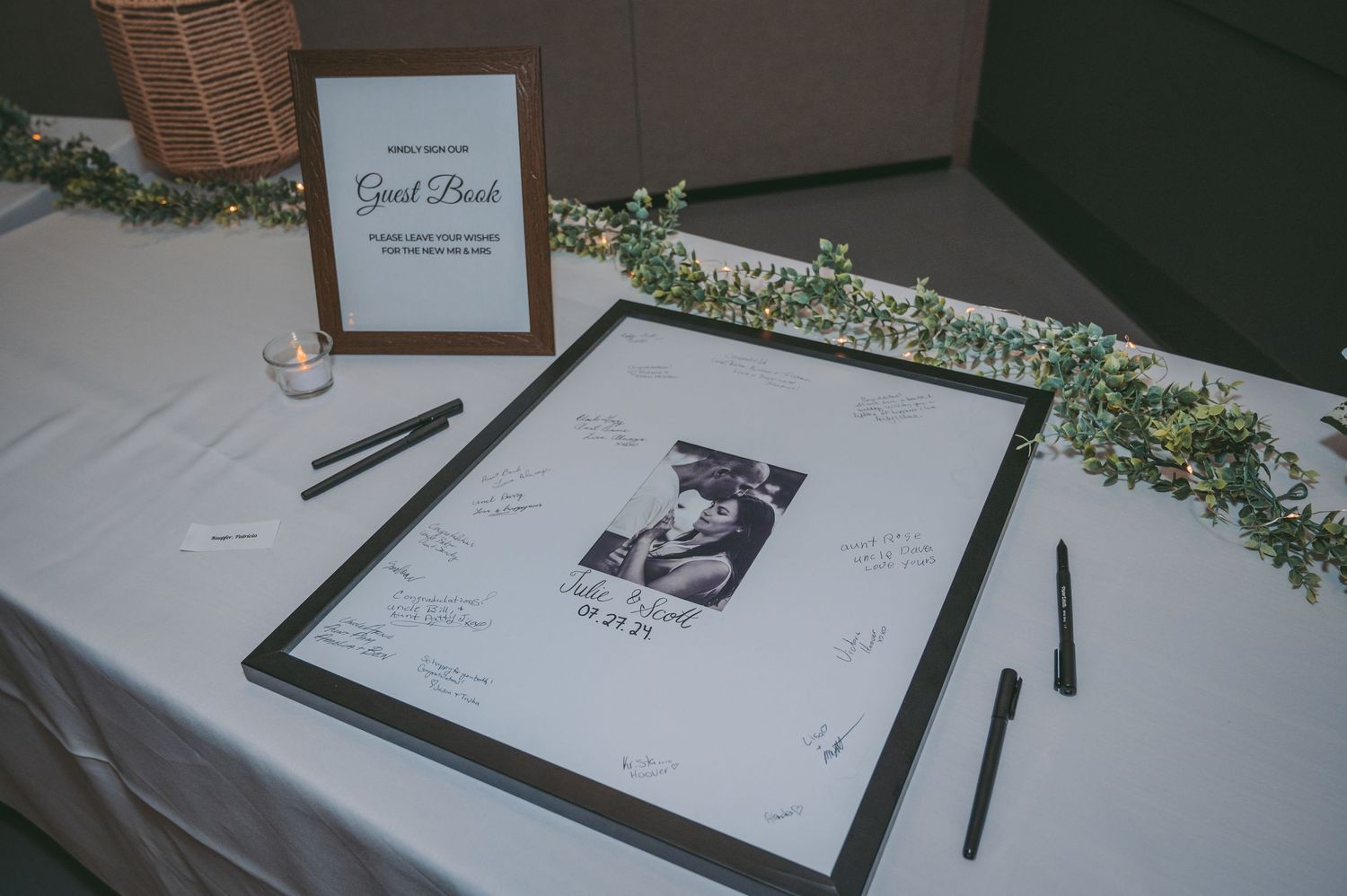 A wedding guest book table setup with framed photo, floral garland, and pens for signing.