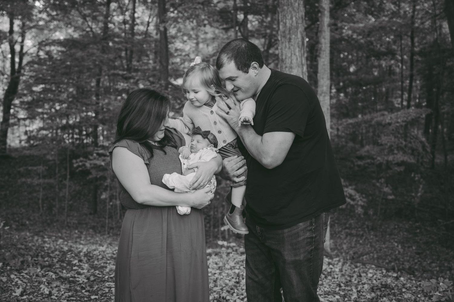 Black and white photo of parents sharing tender moment with children in wooded setting.
