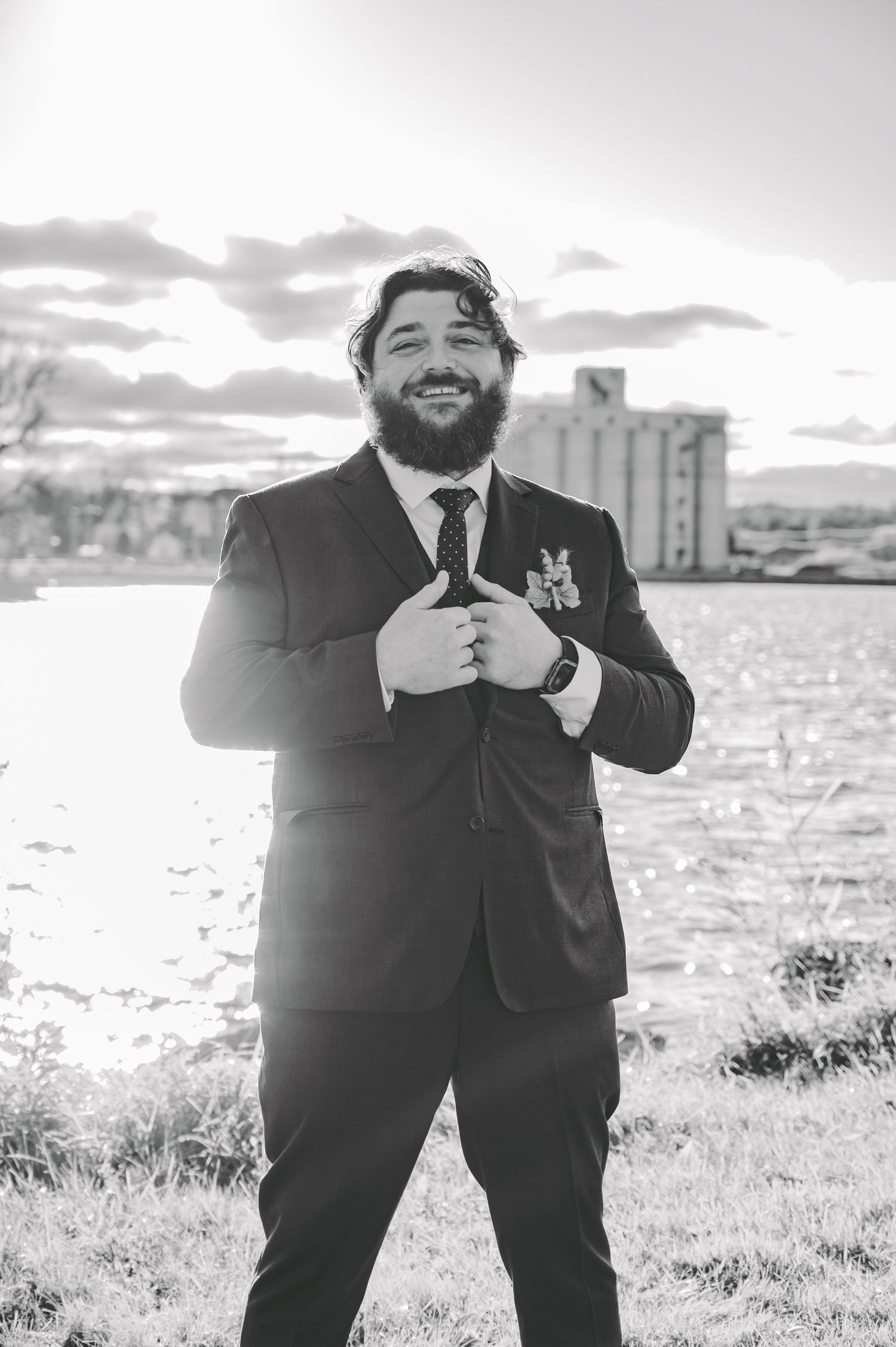 A groom in a classic black suit smiles joyfully during outdoor wedding photos with water and buildings in background.