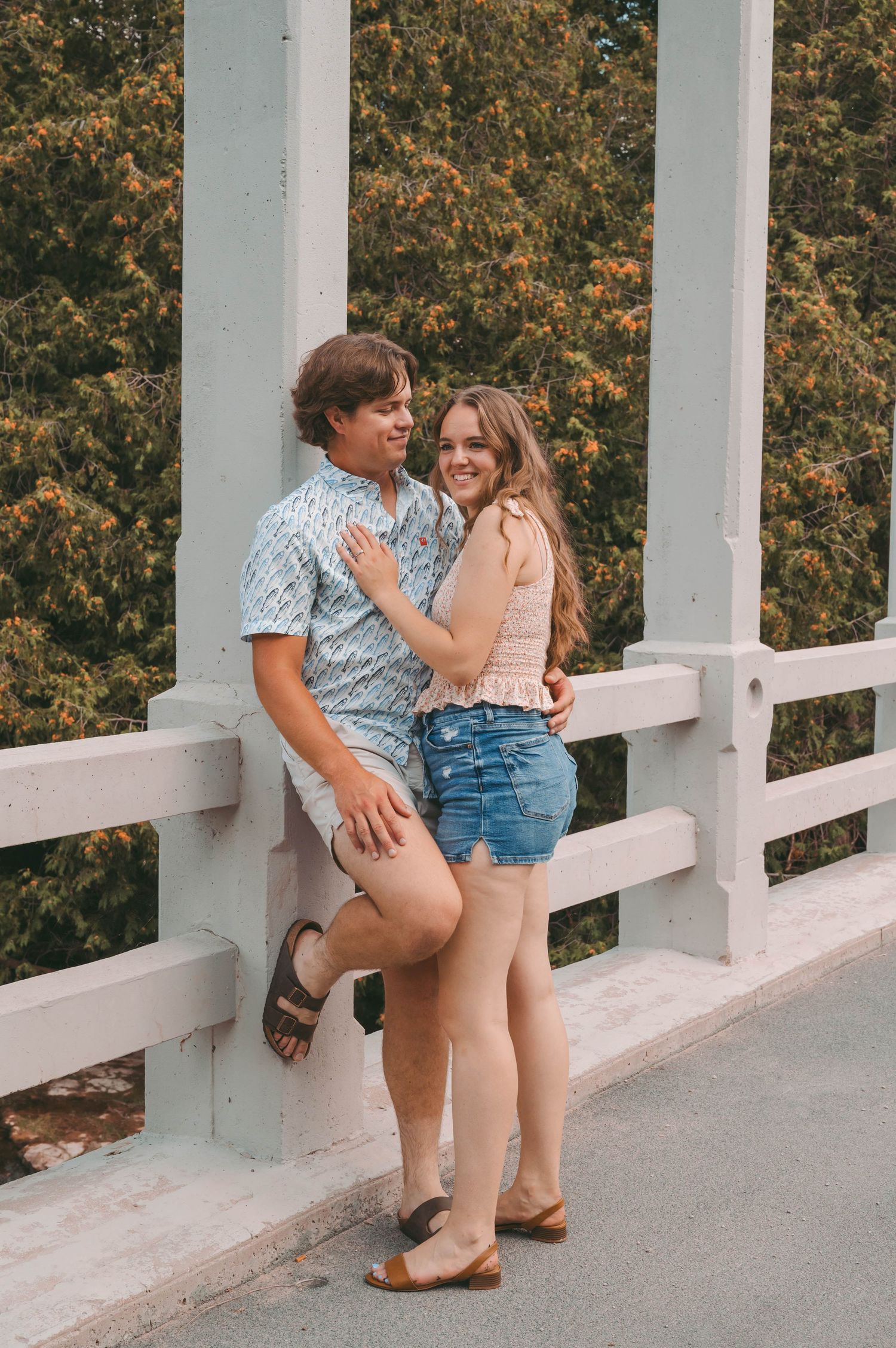 A romantic moment between a couple leaning against a white railing with autumn foliage in the background.