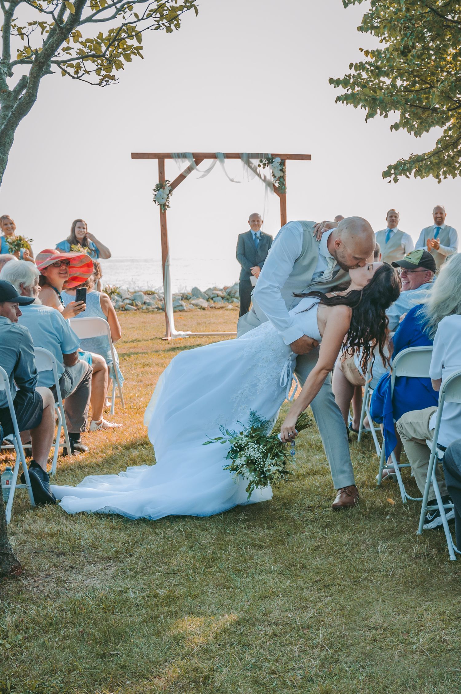 Wedding ceremony dip kiss moment under wooden arch with guests watching by the water.