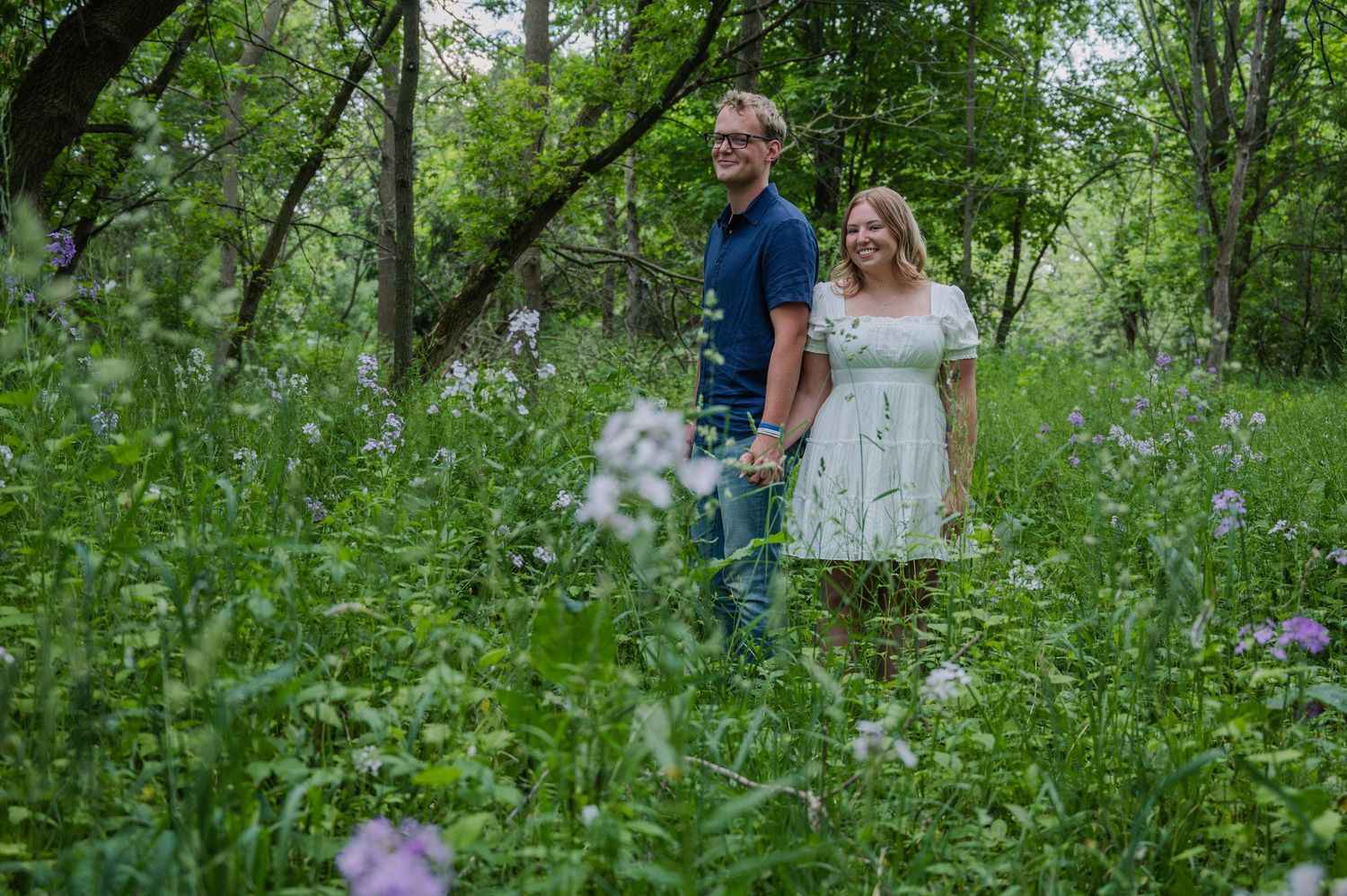Couple holding hands smiling in a lush green wildflower meadow surrounded by trees.