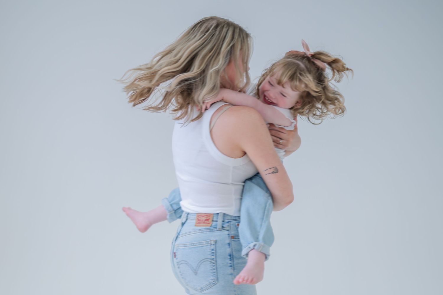 A mother in a white crop top and jeans lovingly holds her young daughter against a light gray background.