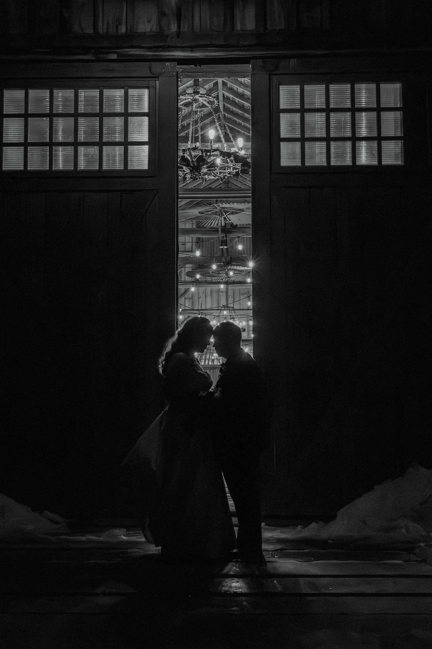 Black and white silhouette of a couple sharing a romantic moment in barn doorway with glowing lights behind them.