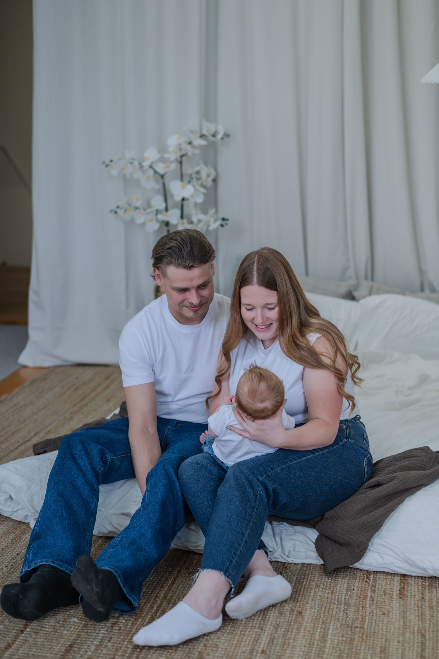 A happy couple sits on a rug holding their newborn baby in a bright, cozy bedroom with white curtains.
