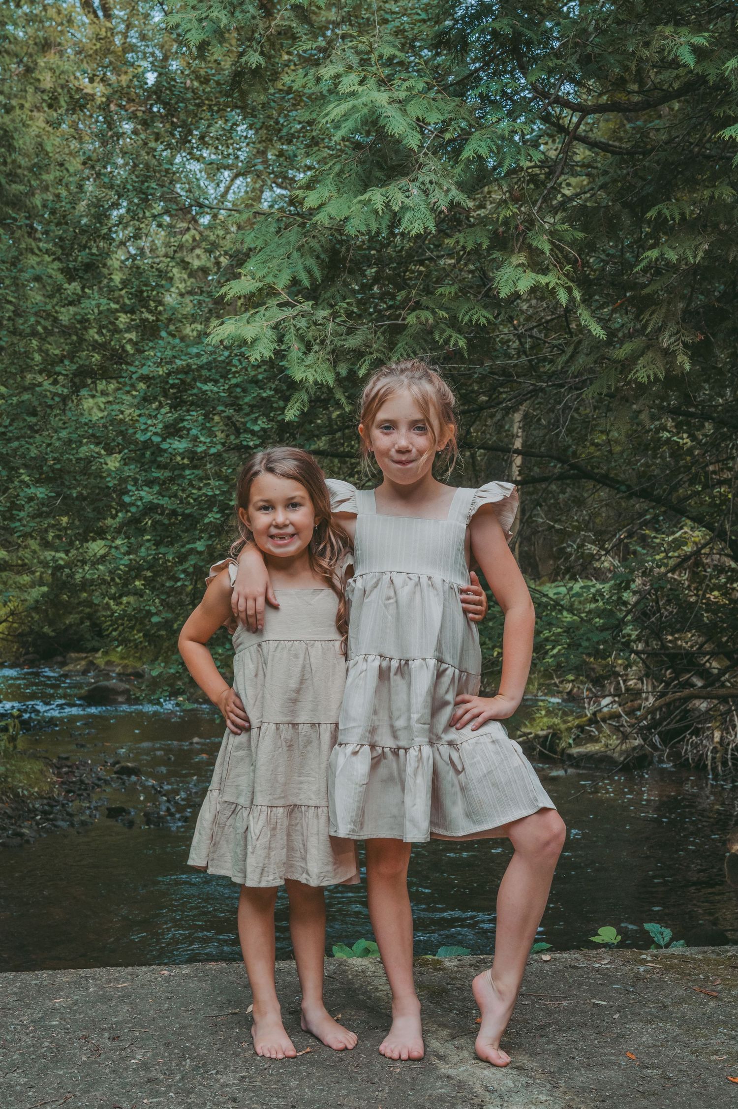 Two young girls in matching dresses pose barefoot by a stream.