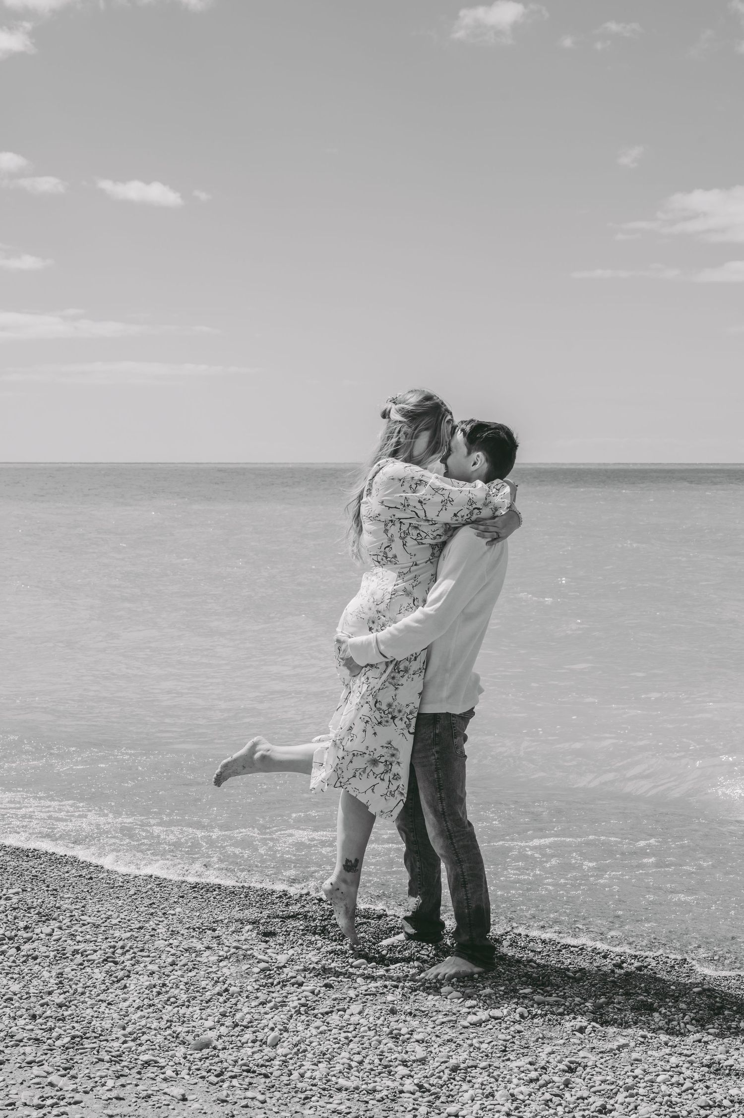 Black and white beach portrait showing playful lift on pebble shore with ocean view.