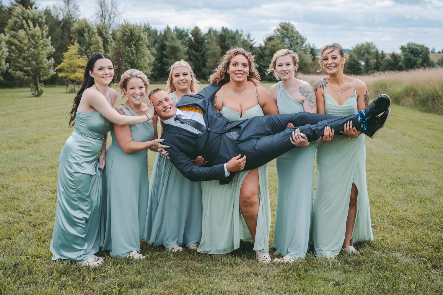 Bridesmaids in mint dresses playfully carrying someone in a fun group photo.
