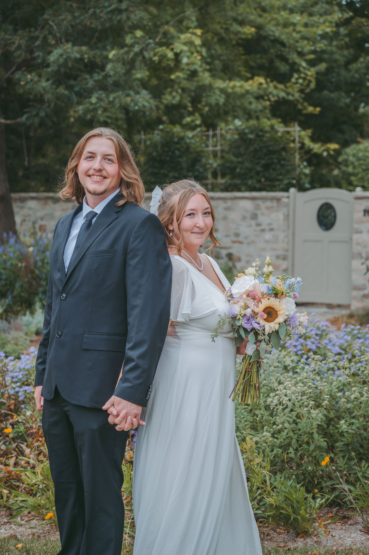 Newlyweds share joyful moments in garden with beautiful summer flowers and stone wall backdrop.