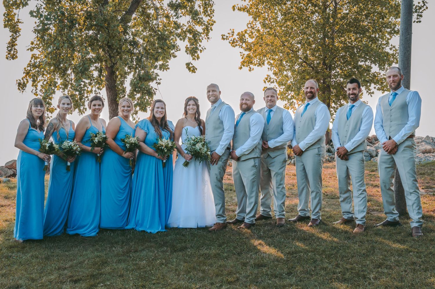 Wedding party in turquoise dresses and light gray suits poses together outdoors.