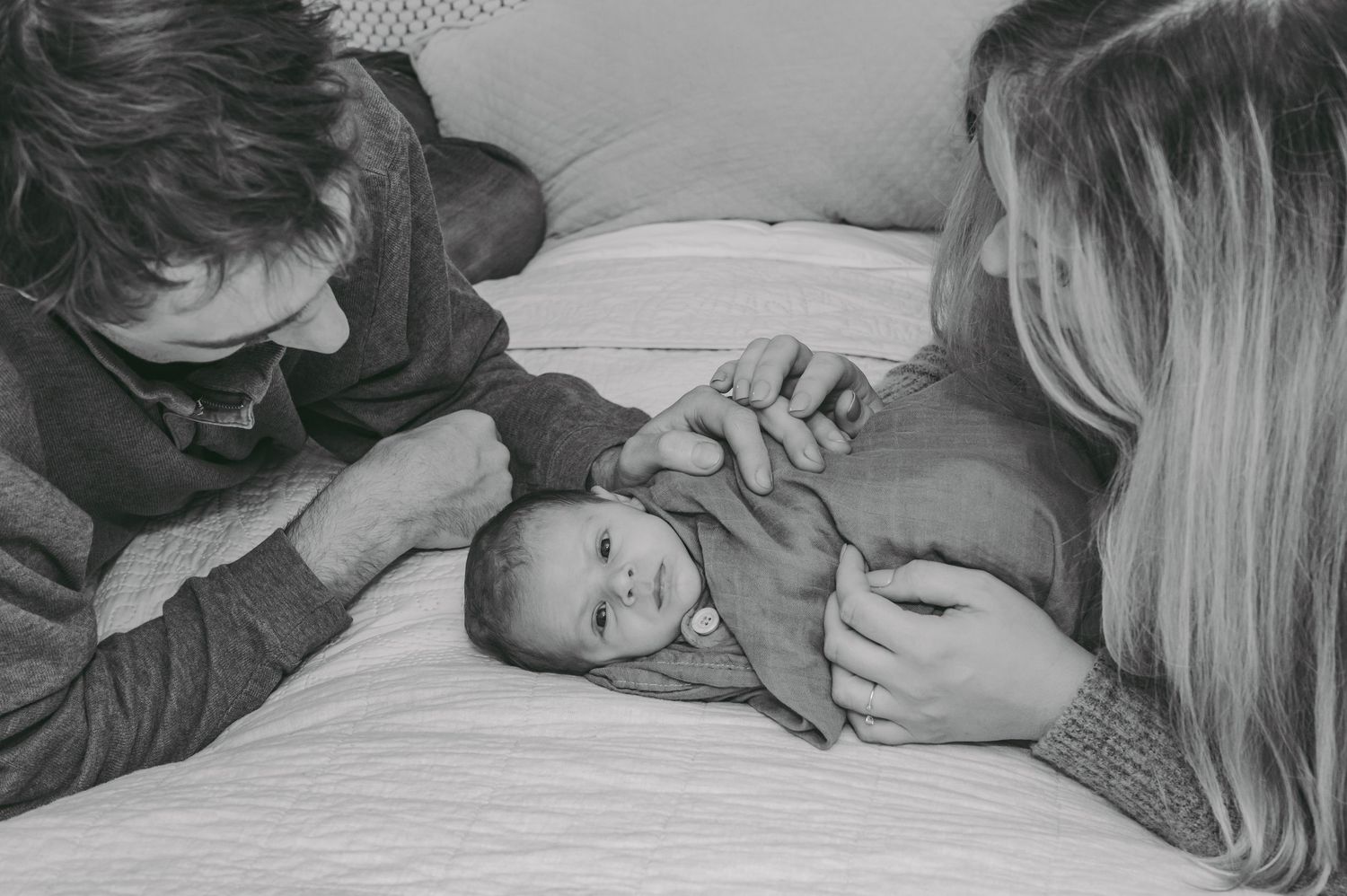 Intimate black and white portrait of parents sharing a tender moment with their newborn on white bedding.