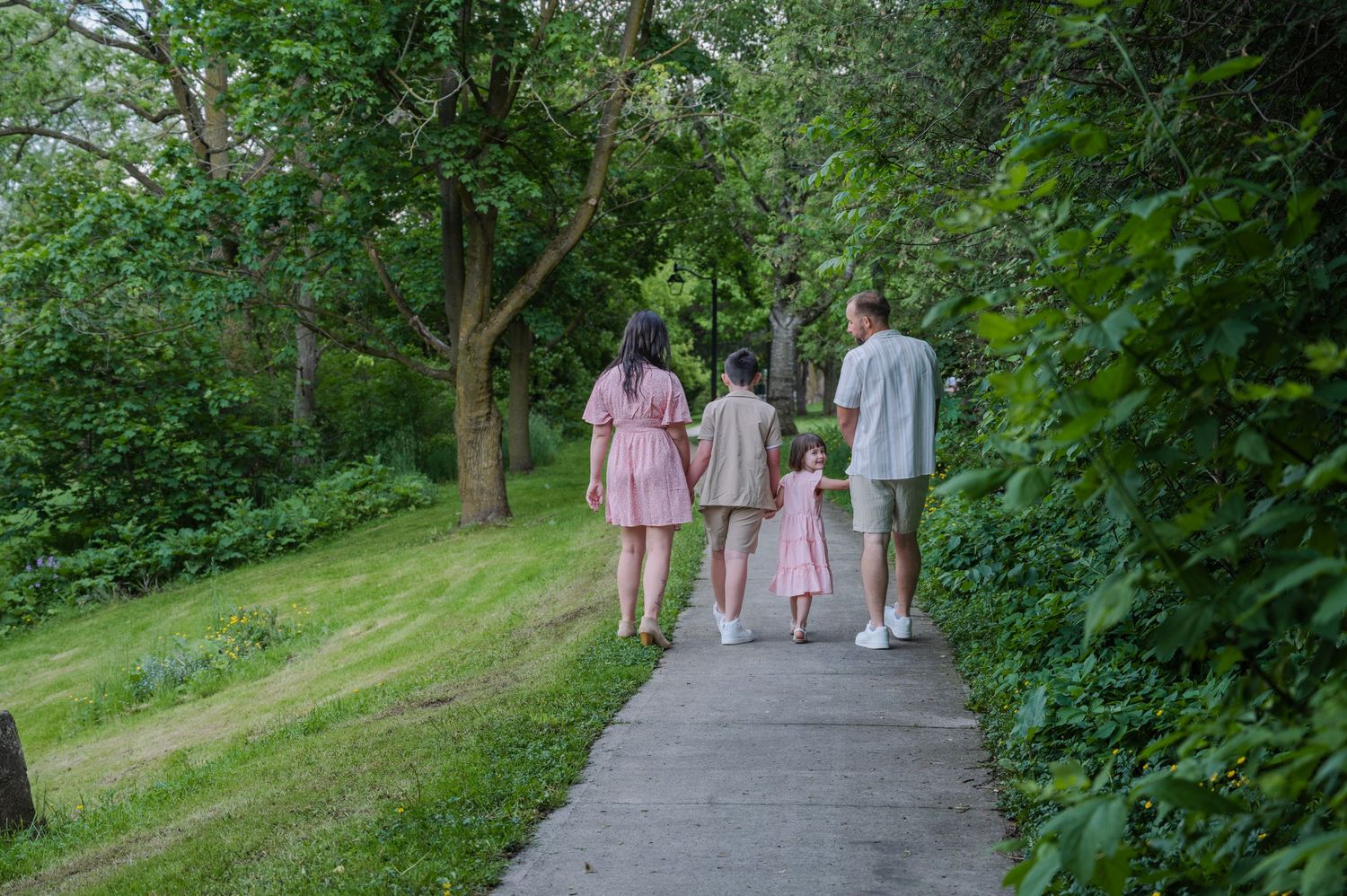 Family of four walking together on a tree-lined path surrounded by lush green trees and grass.