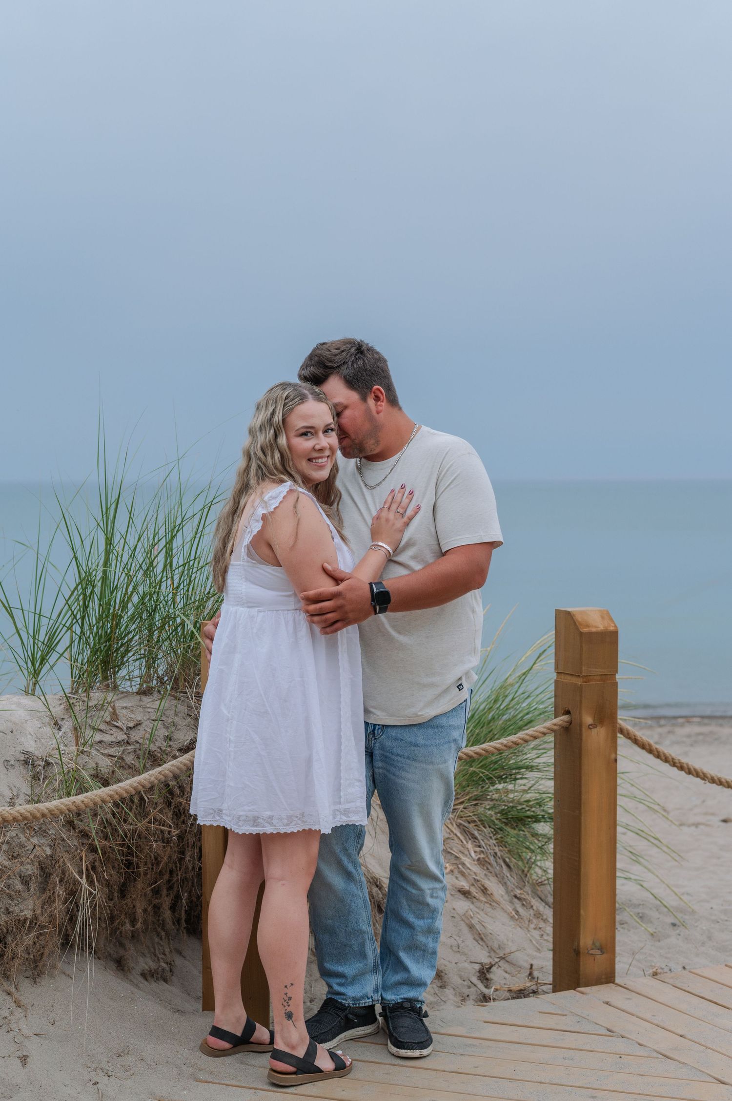 Couple embracing on a sandy beach boardwalk, woman in white dress showing engagement ring, ocean in background.