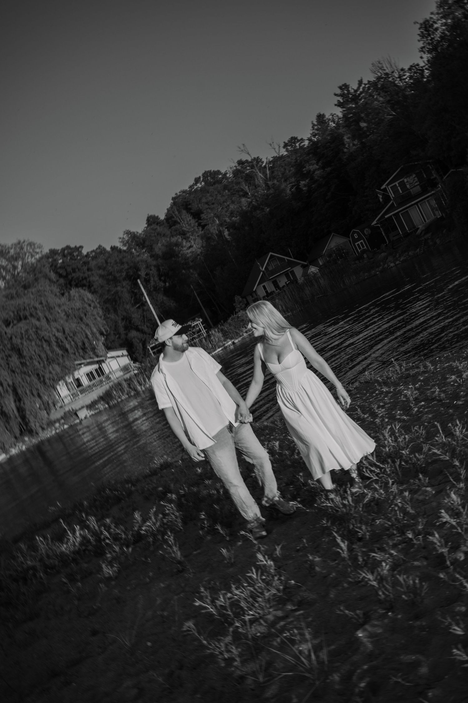 Black and white photo of a couple in white attire holding hands while walking along a lakeside path with trees and buildings behind.