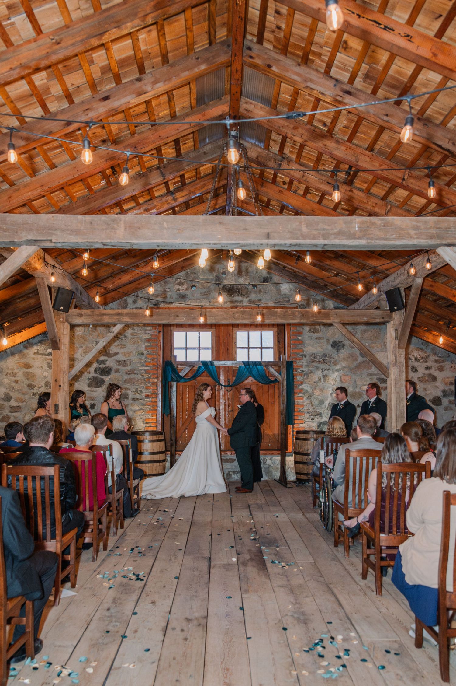 Rustic barn wedding ceremony with string lights, stone walls, and wooden beams as couple exchanges vows at the altar.