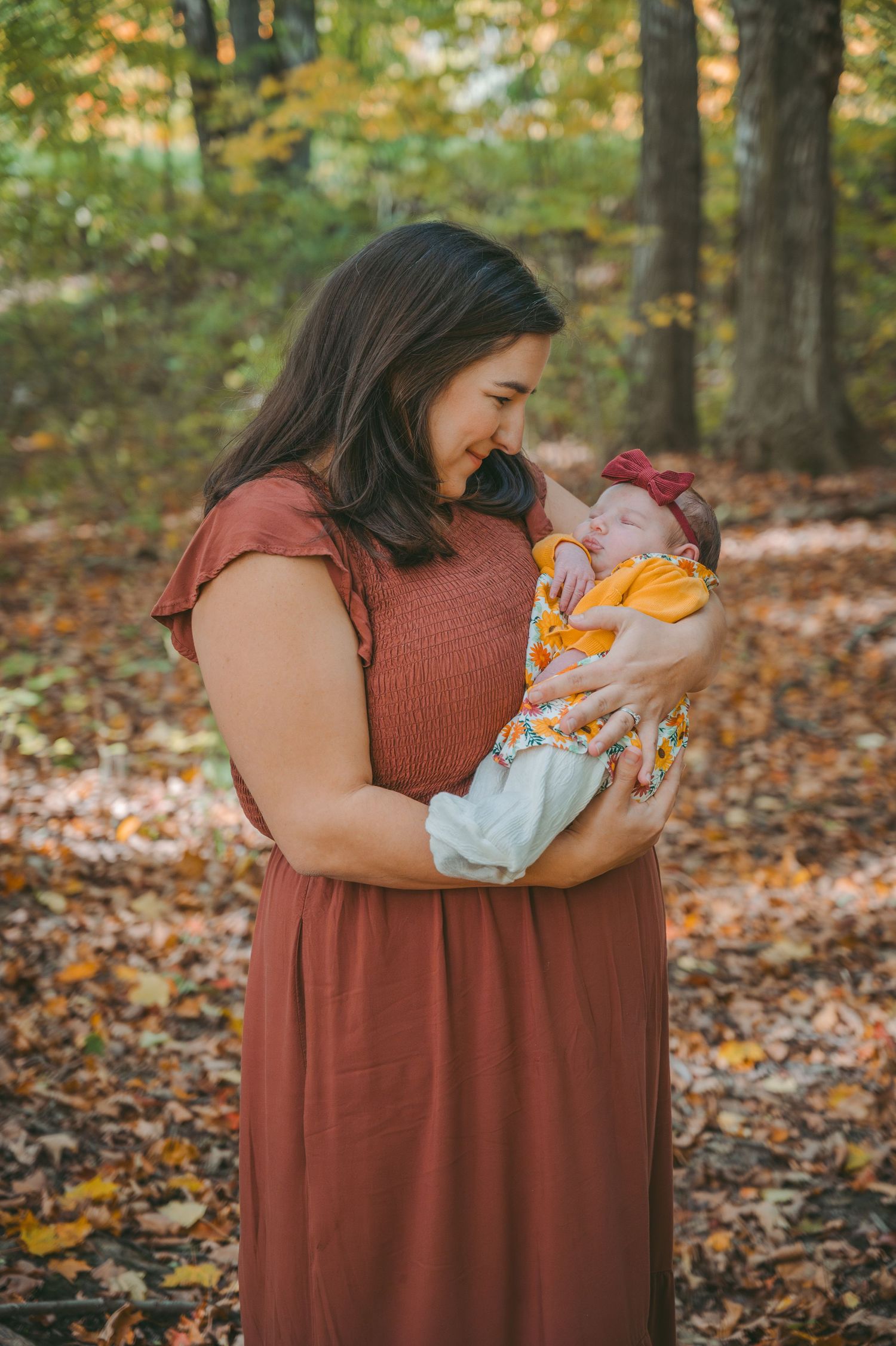Mother in burgundy dress lovingly holds newborn baby against fall forest backdrop.