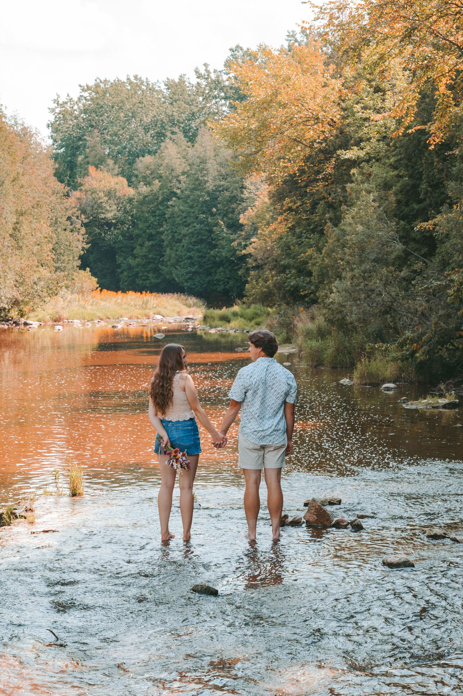 Couple holding hands while standing in riverbed during sunset with fall foliage.