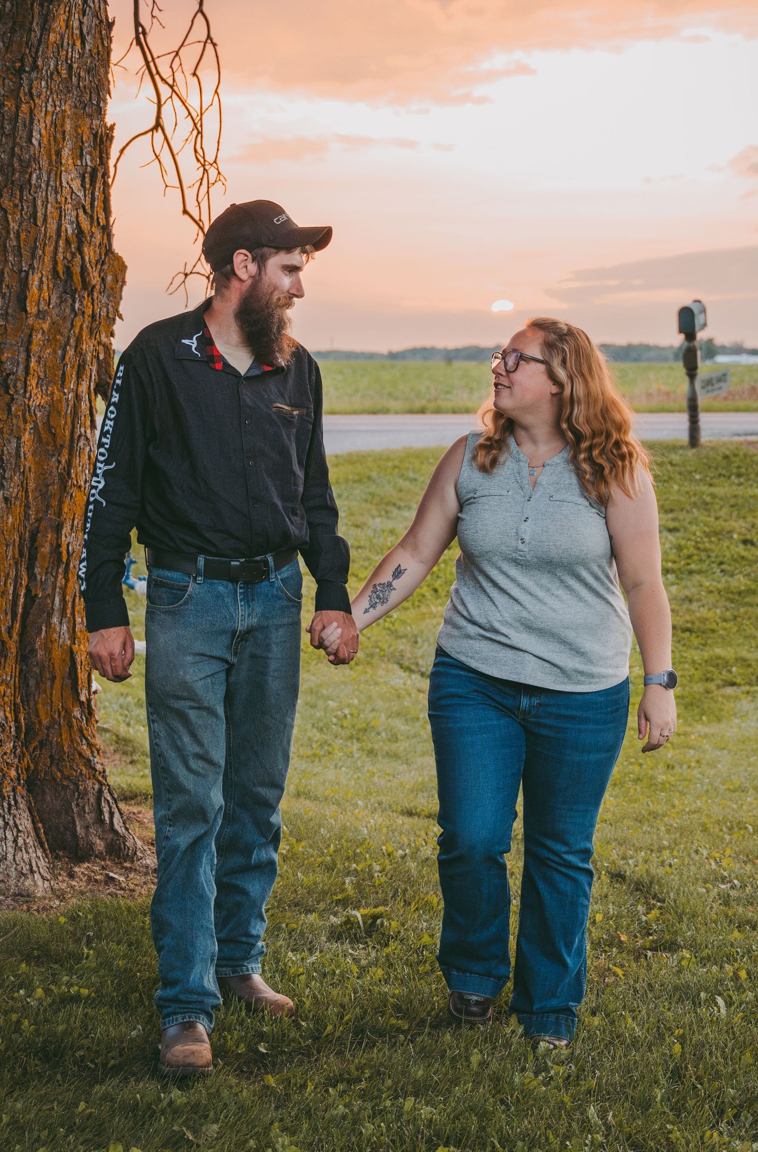 Couple walking hand in hand on a grassy path at sunset beside a large tree.