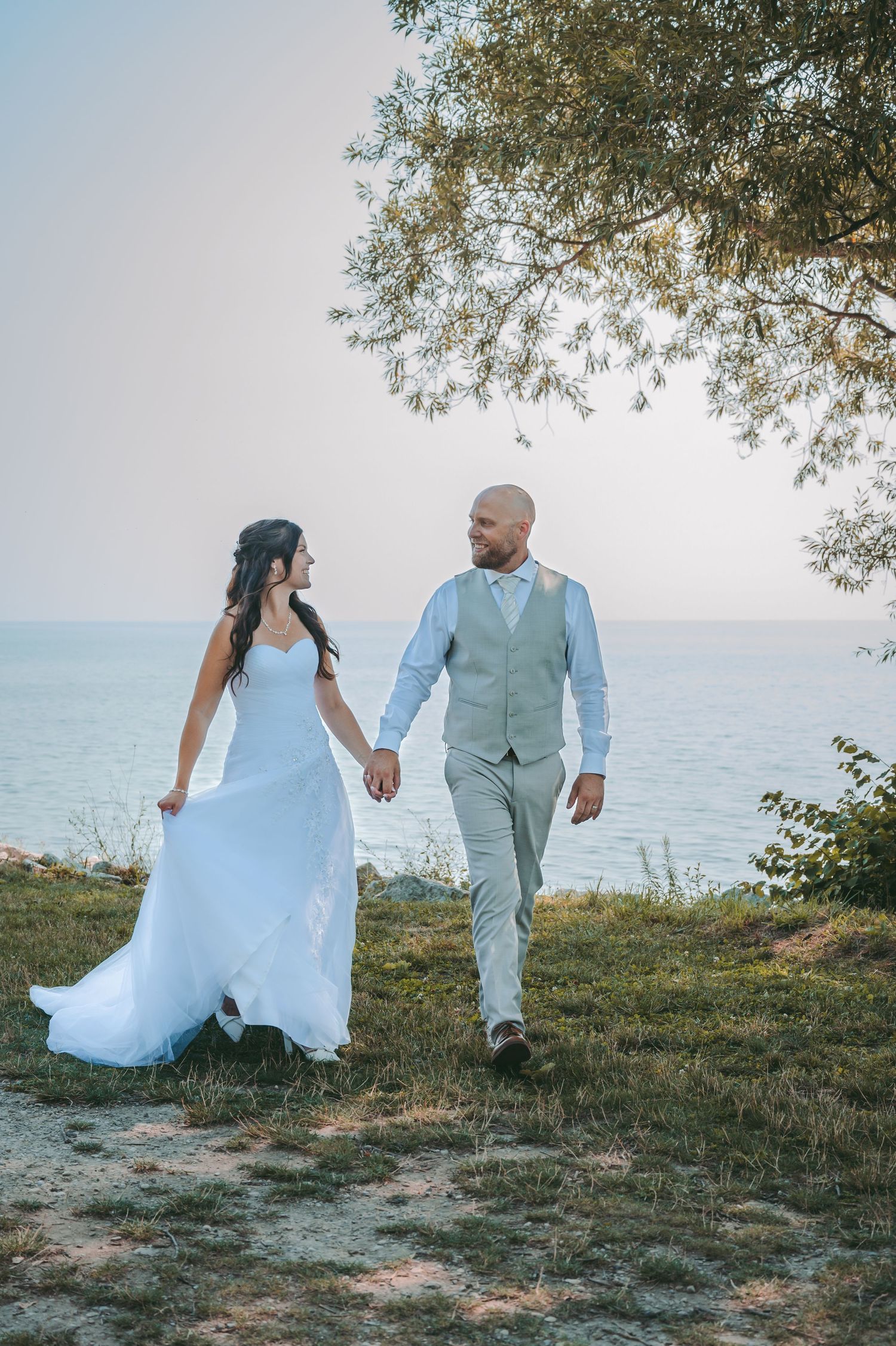 A couple in light colored wedding attire walks along a scenic coastal path during their wedding.