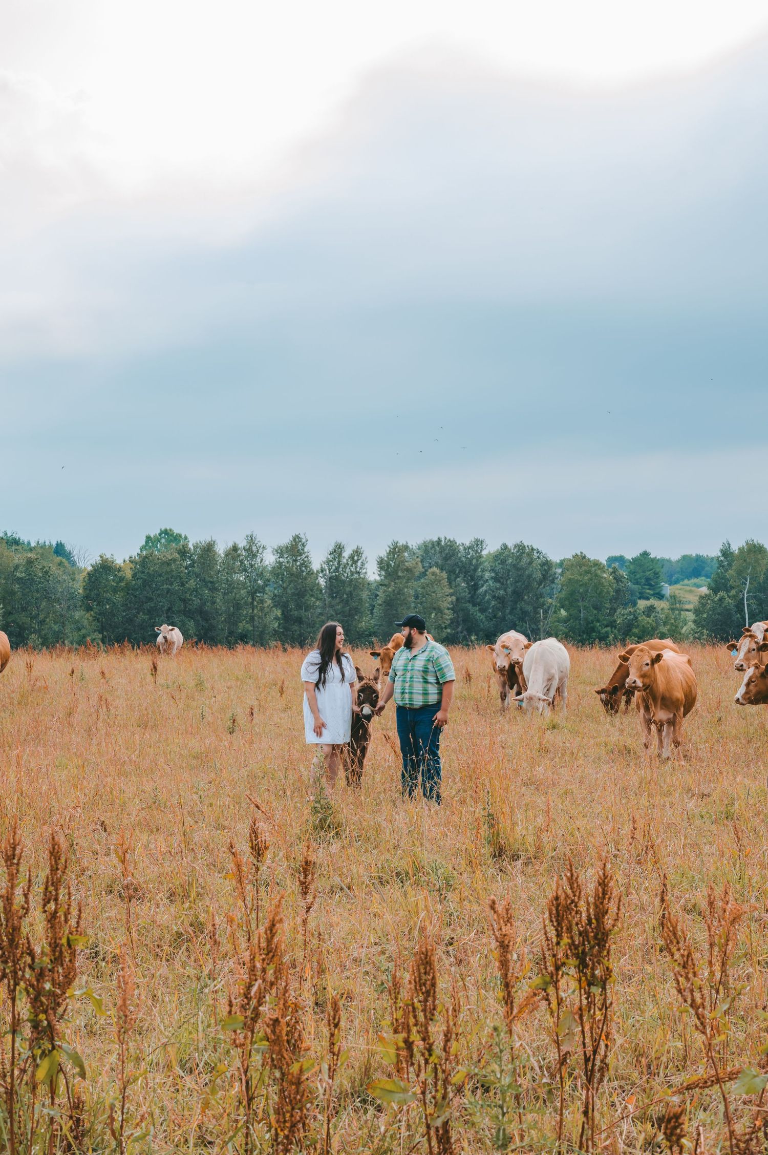 A couple standing in a field with horses grazing in the background on a cloudy day.