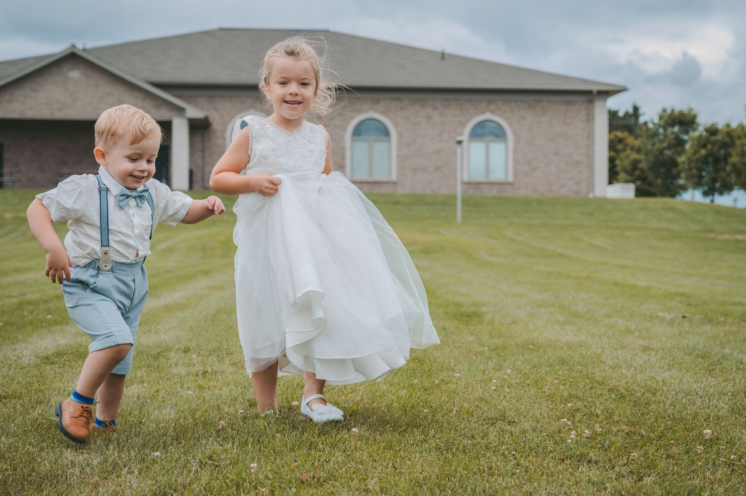 Two children in formal attire running and playing on a grassy lawn.