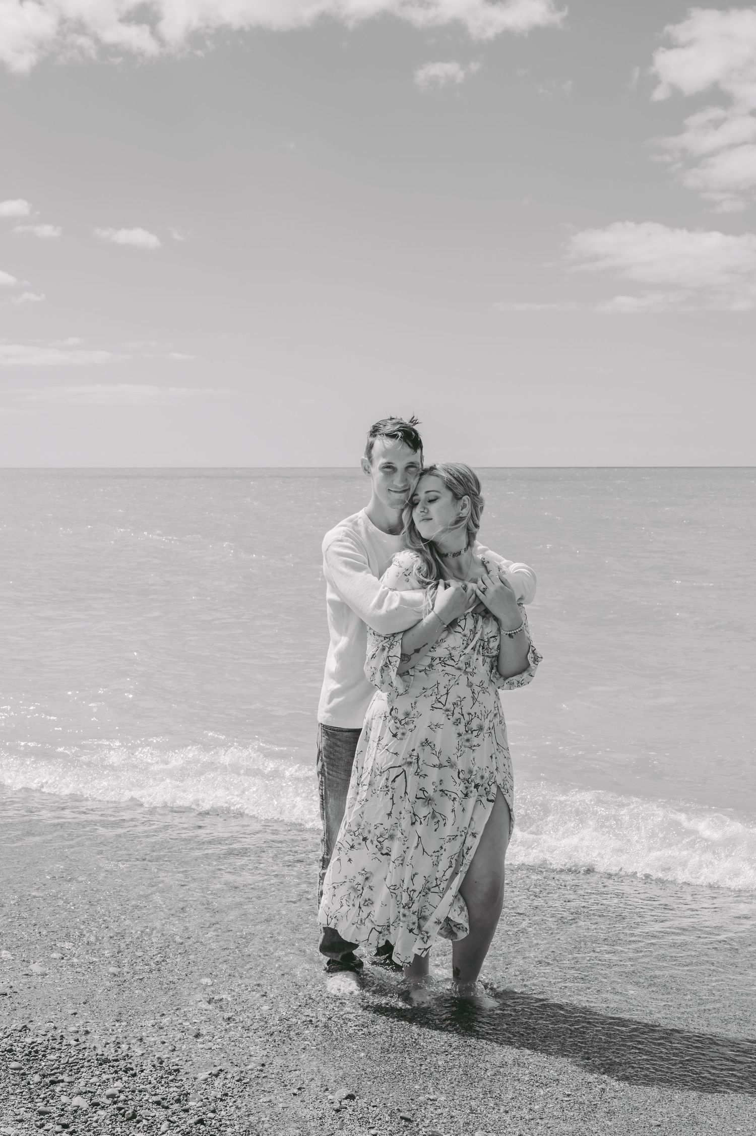Black and white photo of romantic embrace on pebbly beach with waves in background.