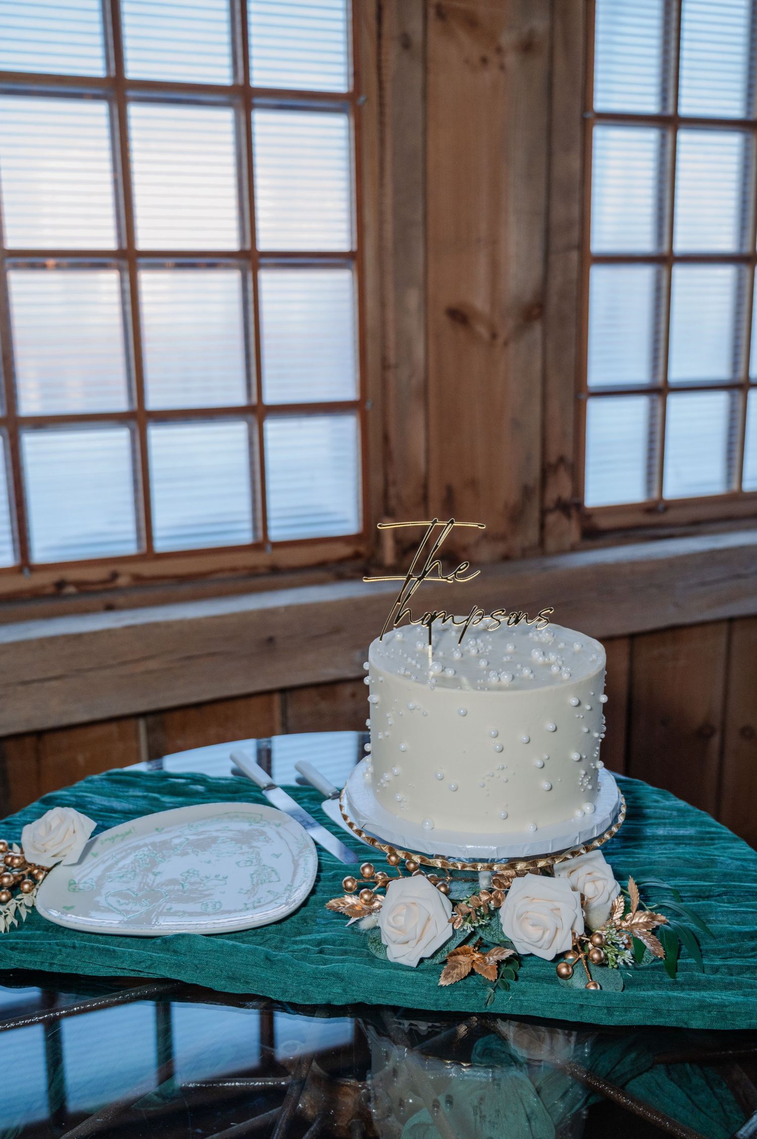 Elegant single-tier white wedding cake with pearl details and gold topper on a teal table with roses.