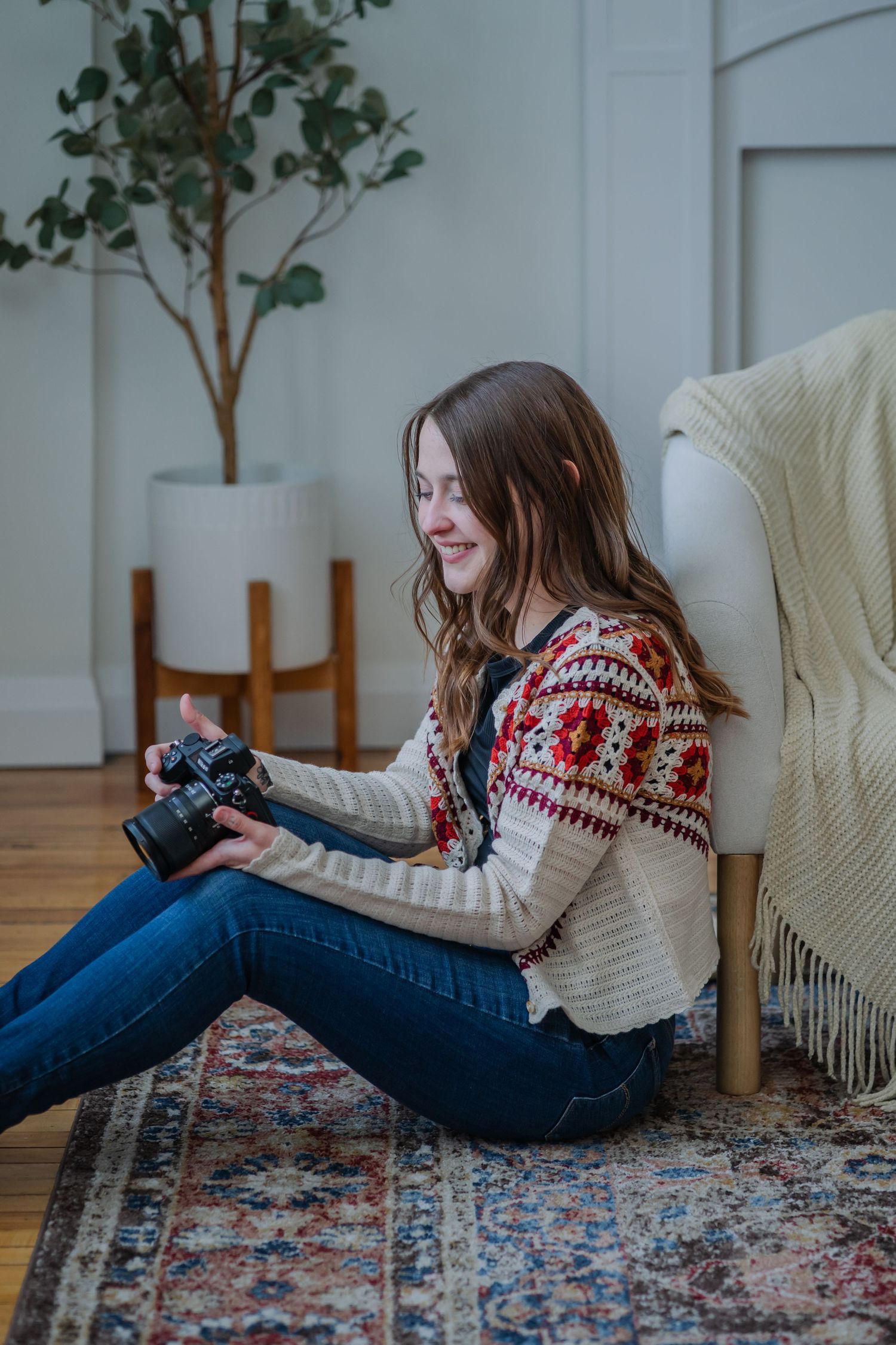 Person in red patterned sweater and jeans lounges on ornate rug while using camera in bright room with plants.