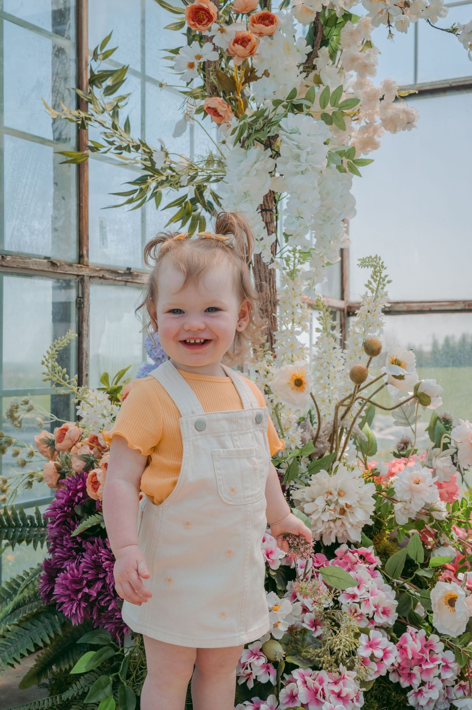 A toddler in white overalls and yellow top plays among colorful flowers in a greenhouse setting.