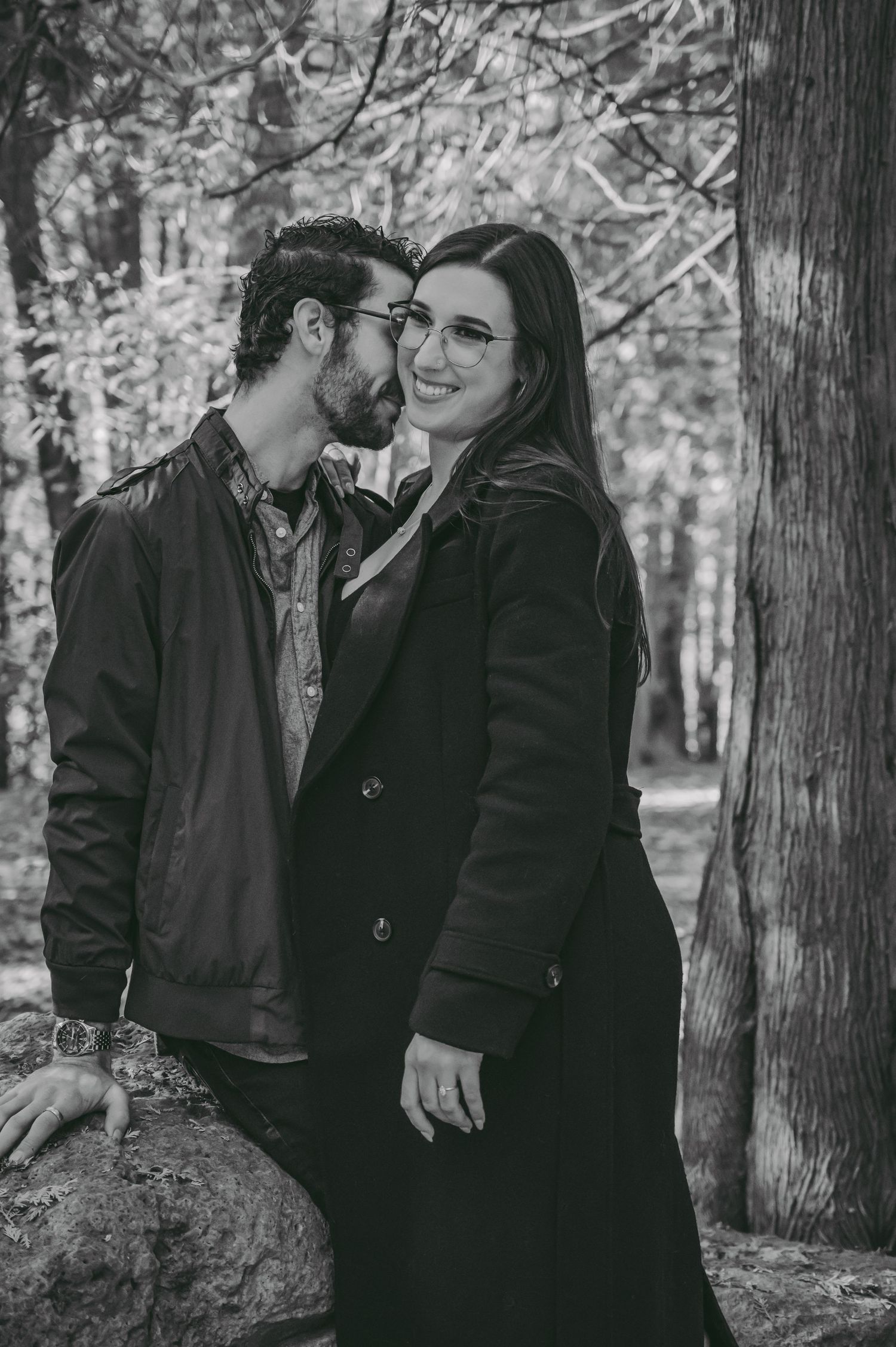 A couple shares an intimate moment against a forest backdrop in black and white photography.