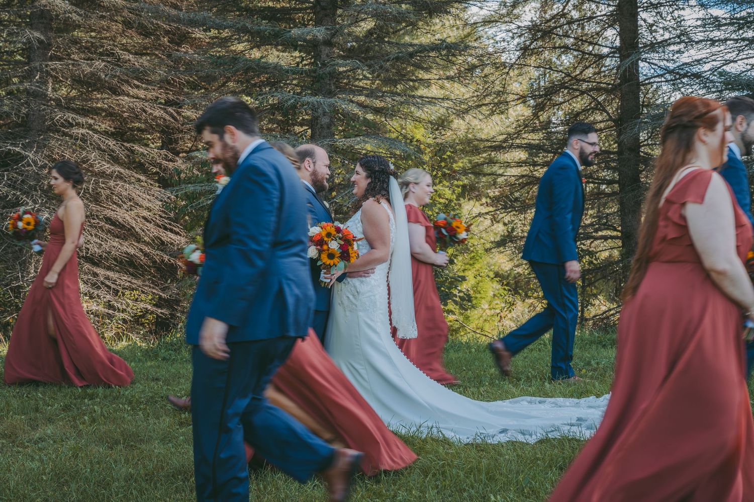 Wedding party walking through a forest setting with the bride in white and attendants in burgundy and navy.