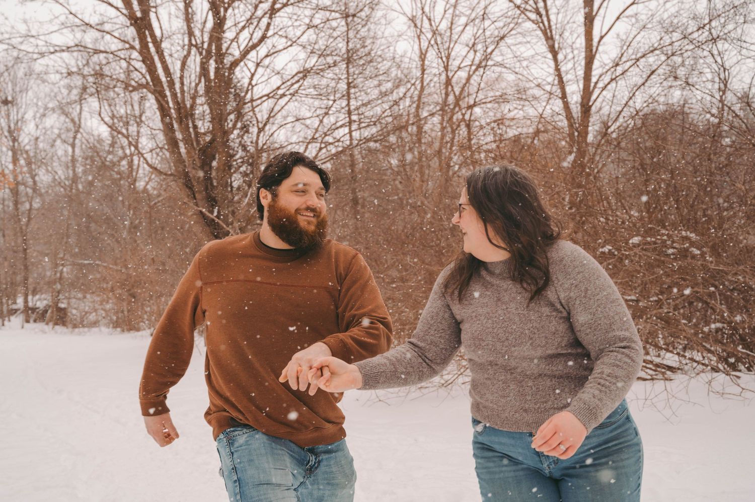 Couple walking hand in hand through snowy landscape wearing casual winter attire.