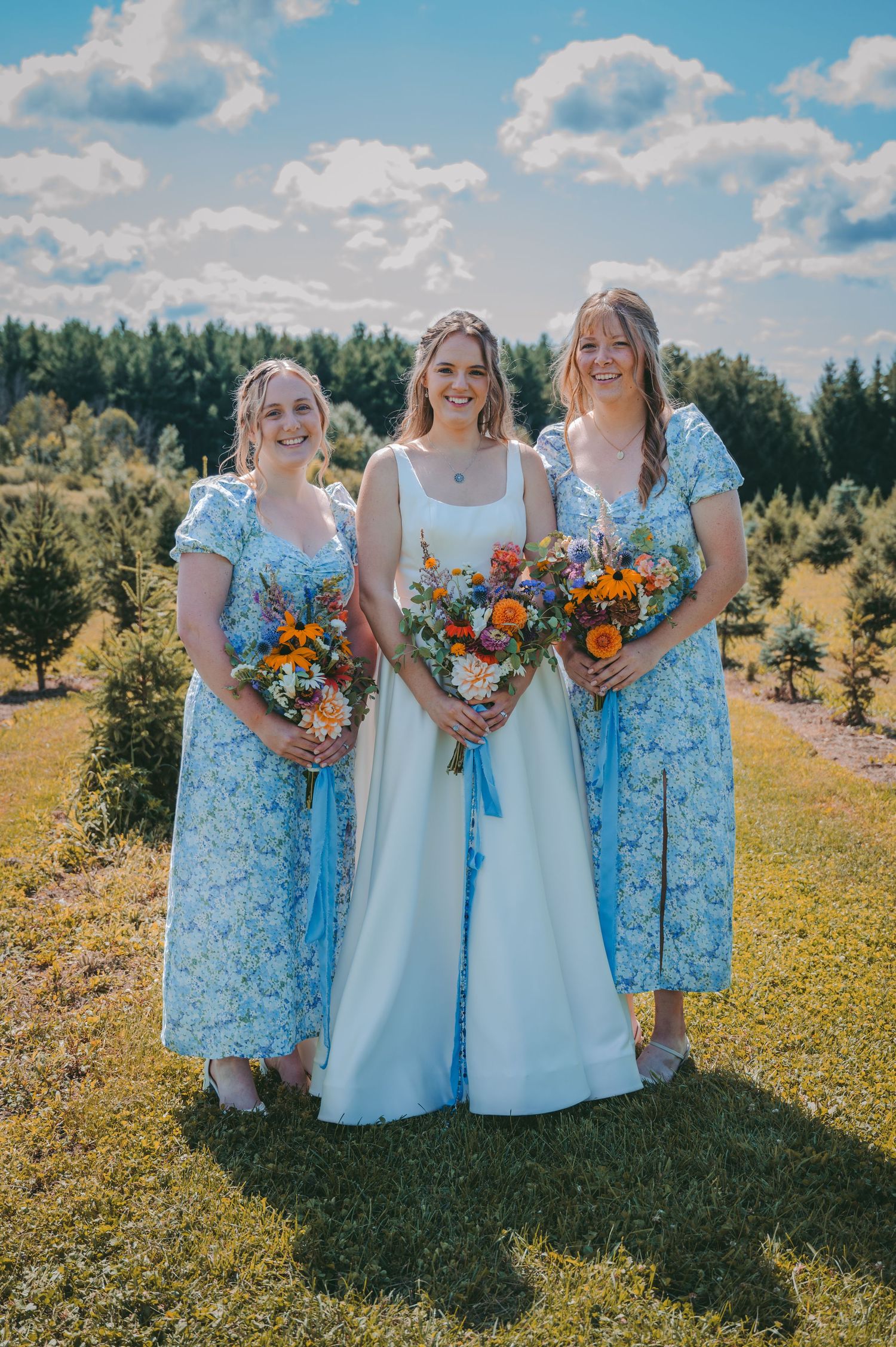 Bridesmaids in light blue floral dresses pose with vibrant orange and white bouquets in a sunny outdoor field.