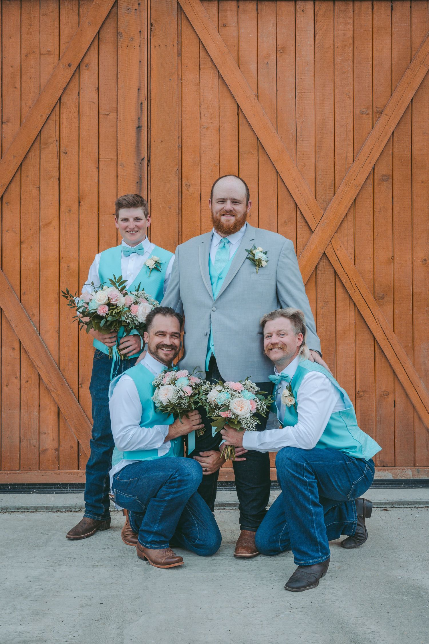 Wedding party members in light blue shirts and navy pants kneel in front of rustic wooden barn doors.