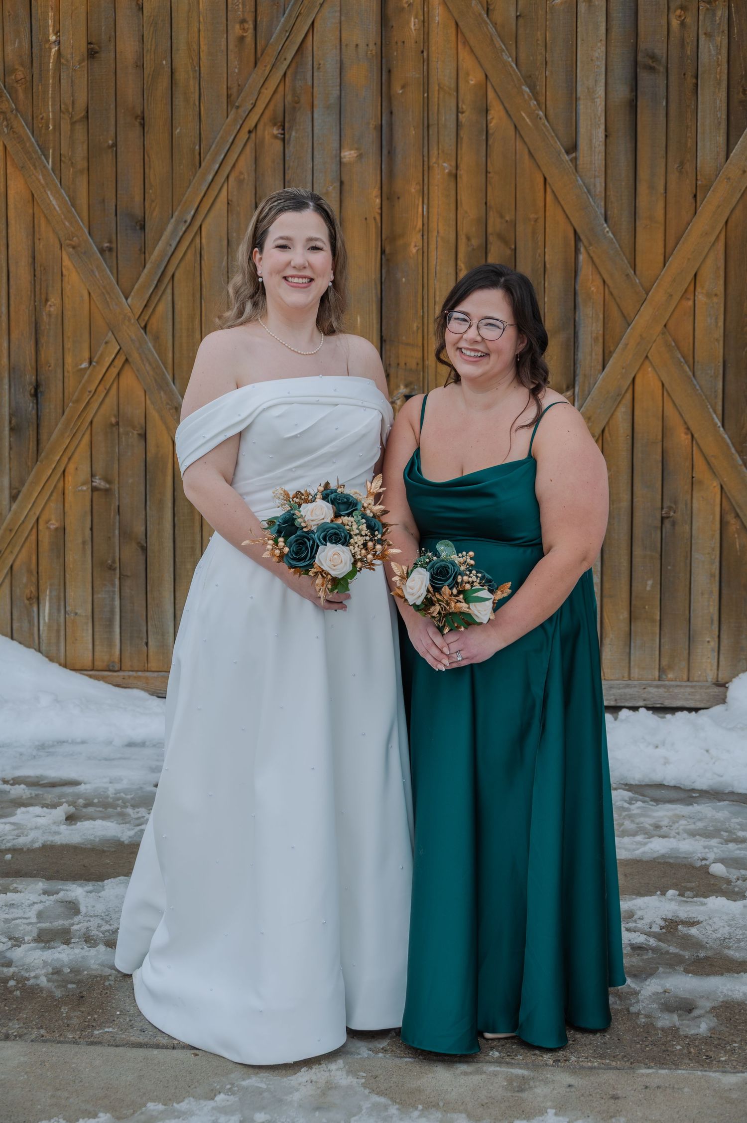 Bride in white off-shoulder gown and bridesmaid in emerald green dress holding bouquets before rustic barn doors.