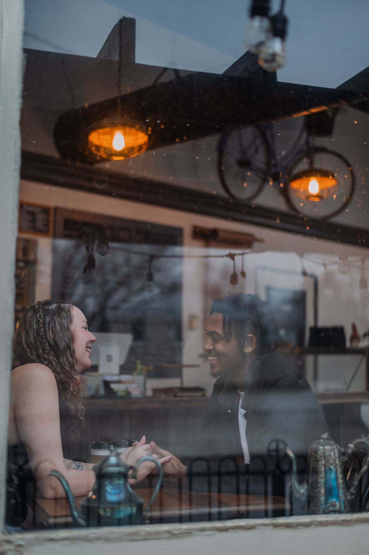 Couple laughing together through a cafe window, warm Edison bulbs glowing above, bicycle visible in background.