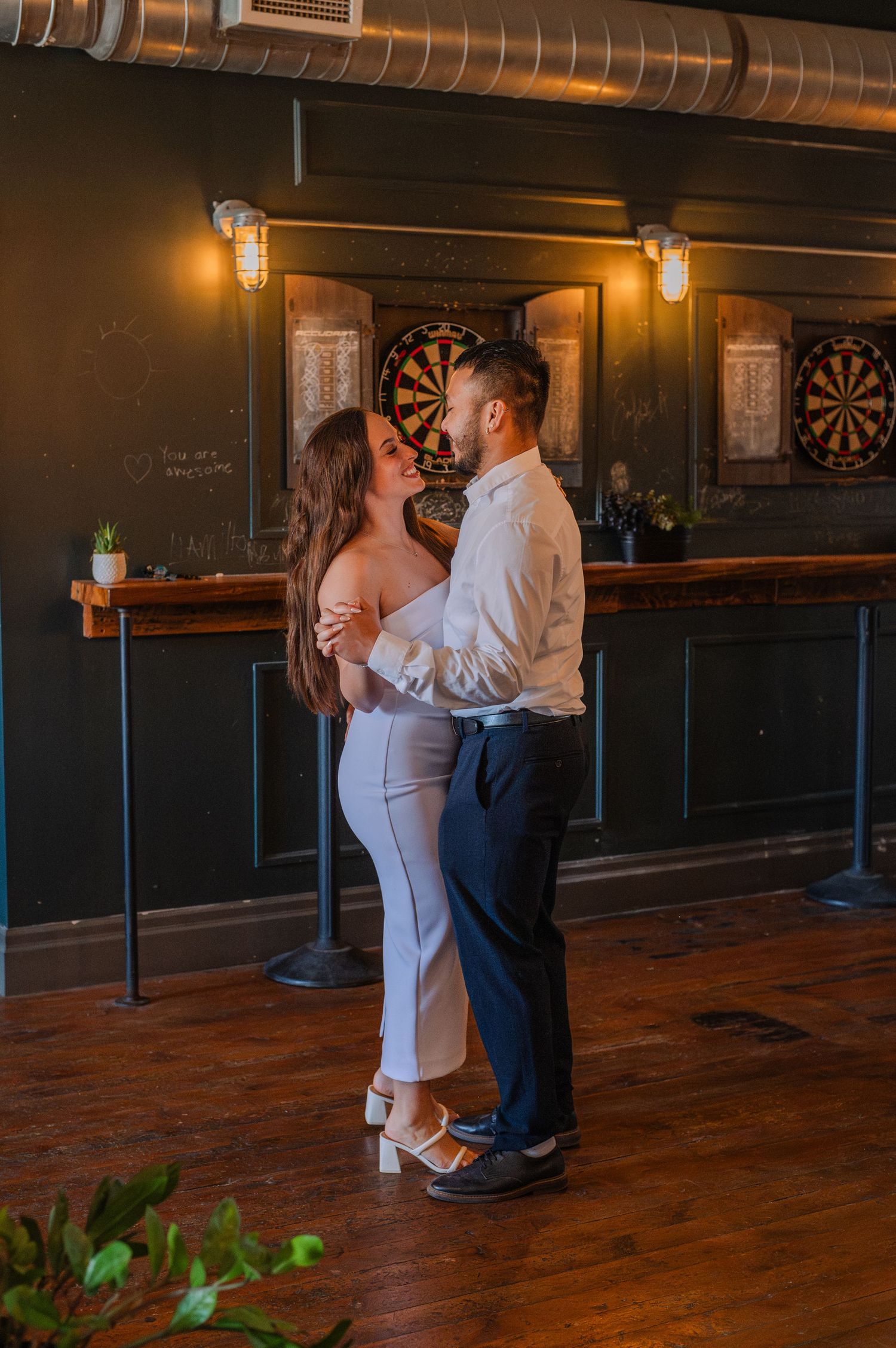 Couple slow dancing in a dimly lit bar with dartboards and warm lighting in the background.