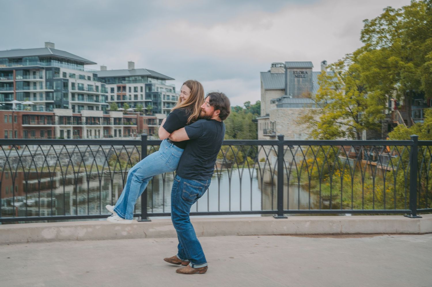 Playful couple dancing together on waterfront plaza with city buildings in background.
