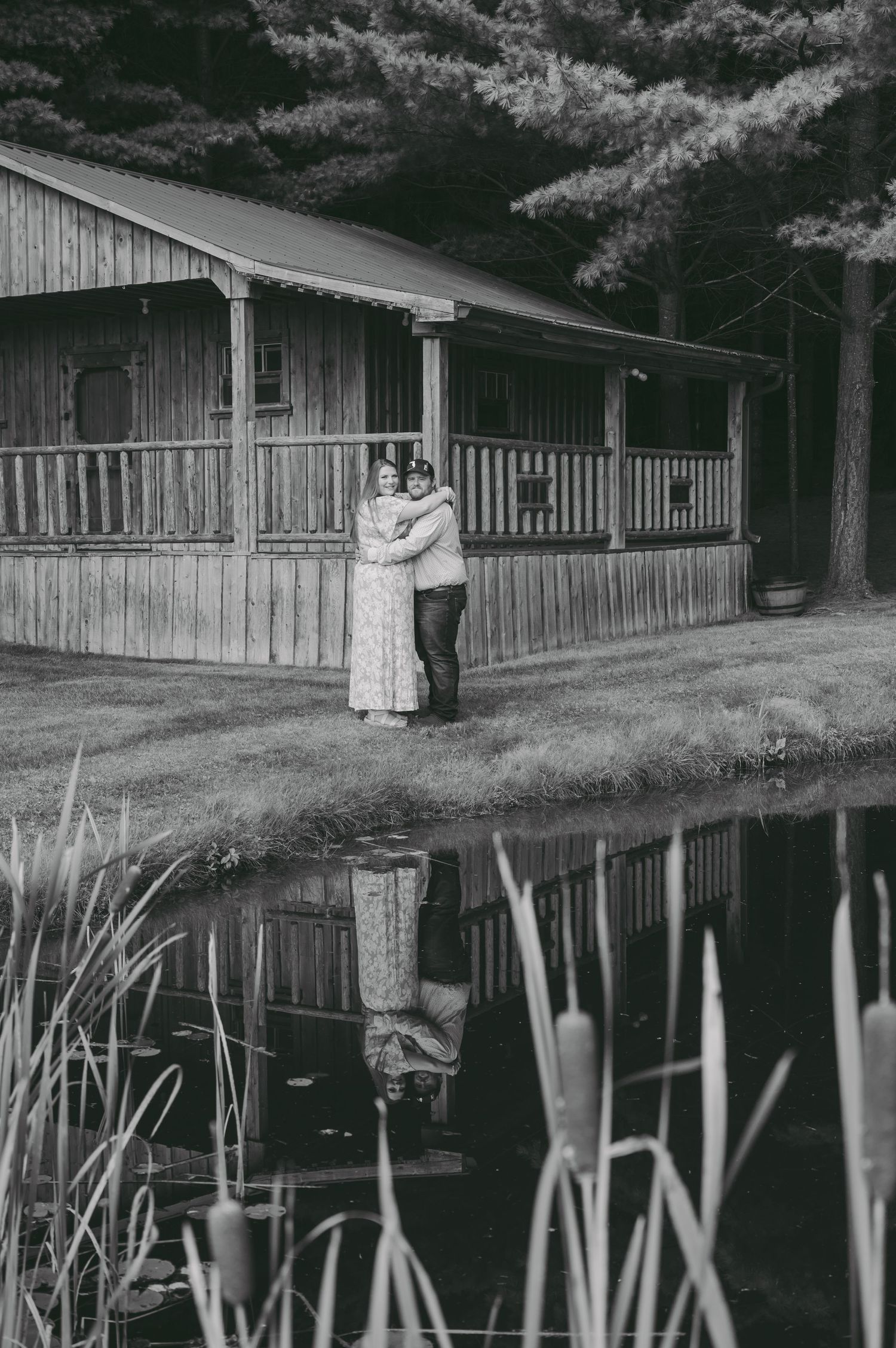 A couple embraces in front of a rustic wooden cabin with a pond reflection in black and white.