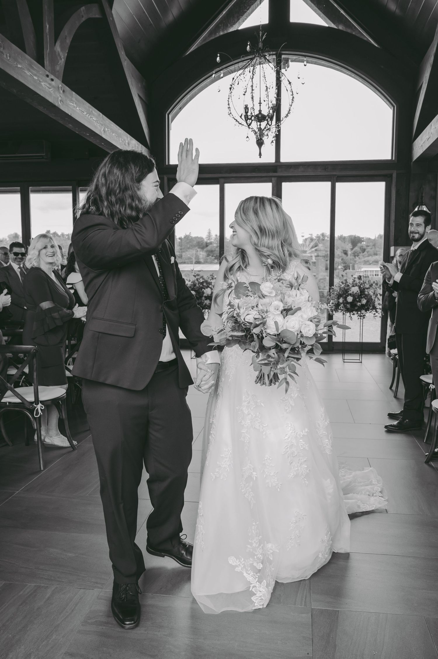 Black and white photo of wedding celebration moment under elegant chandelier and vaulted ceiling.