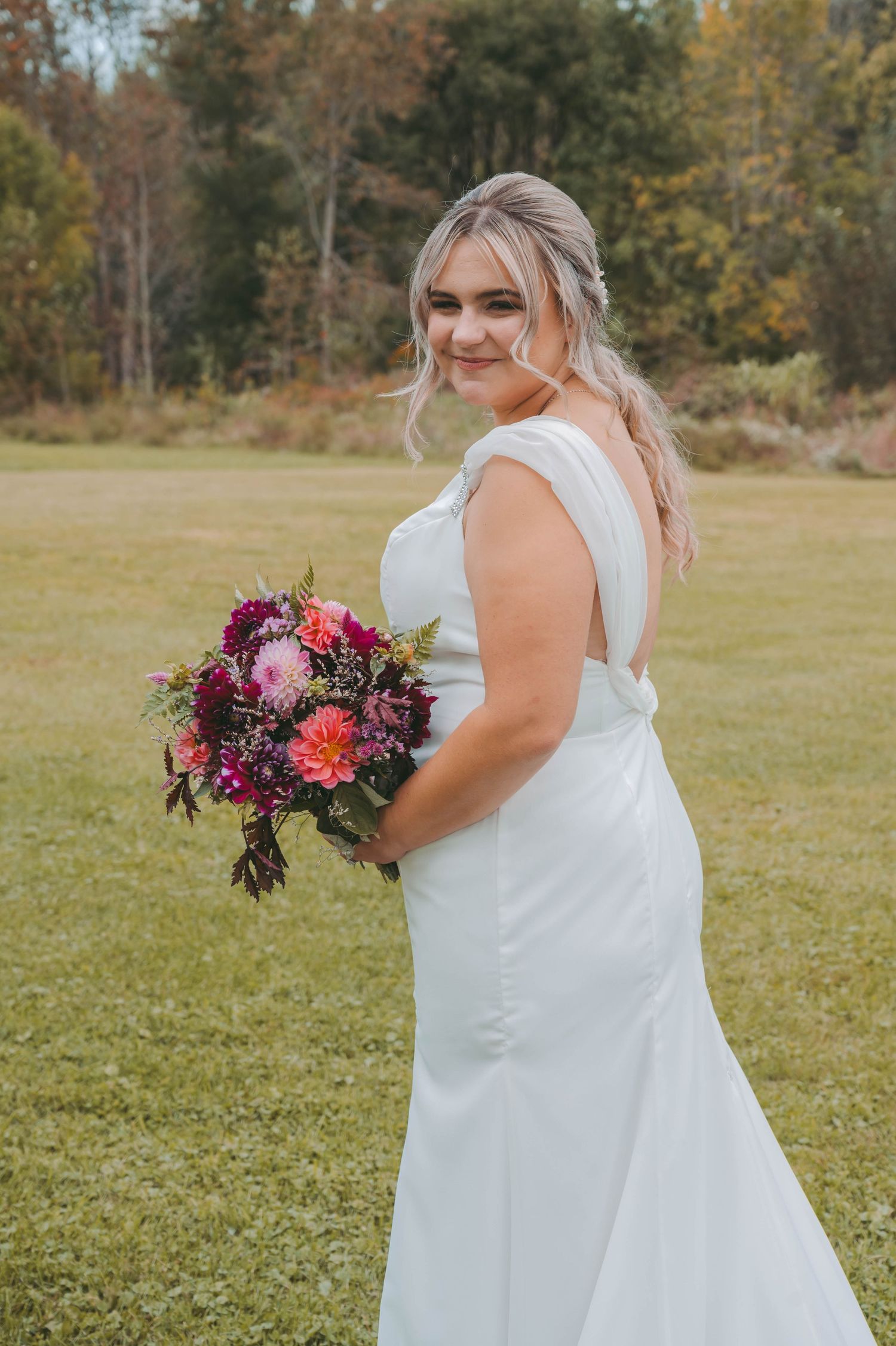 Bride in white dress holding vibrant purple and pink bouquet in outdoor setting.