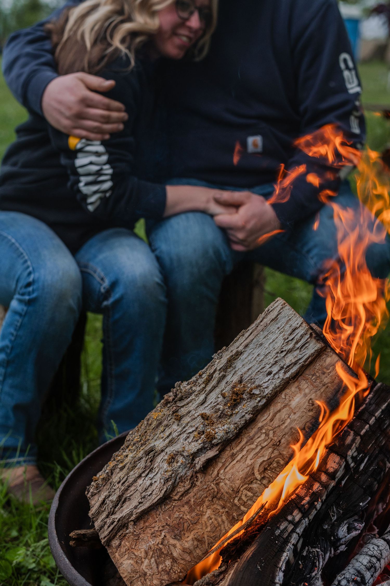 Two people in Carhartt hoodies sit together near a crackling wood fire pit outdoors.