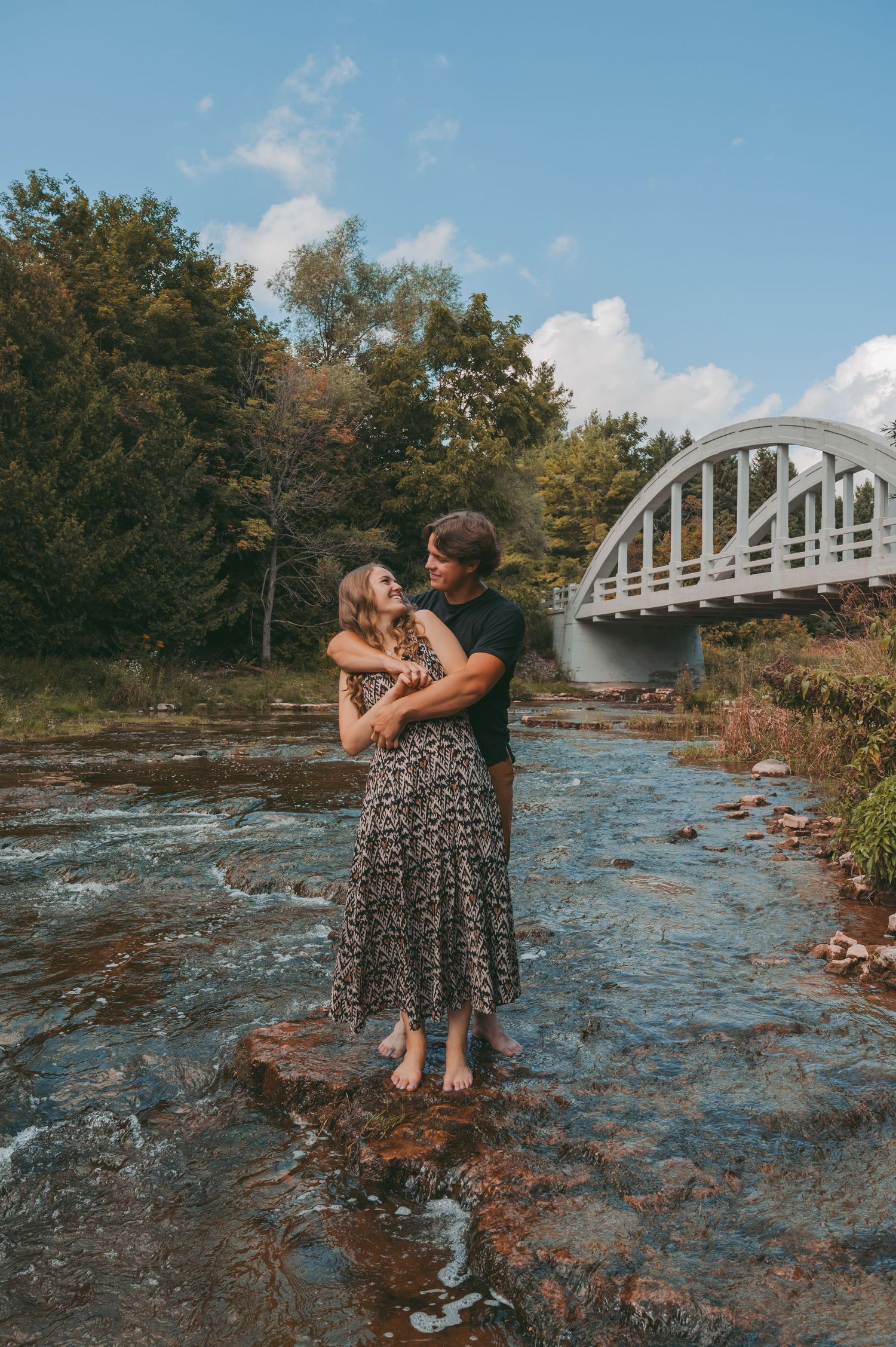Romantic couple embracing in shallow river water beneath historic white bridge.